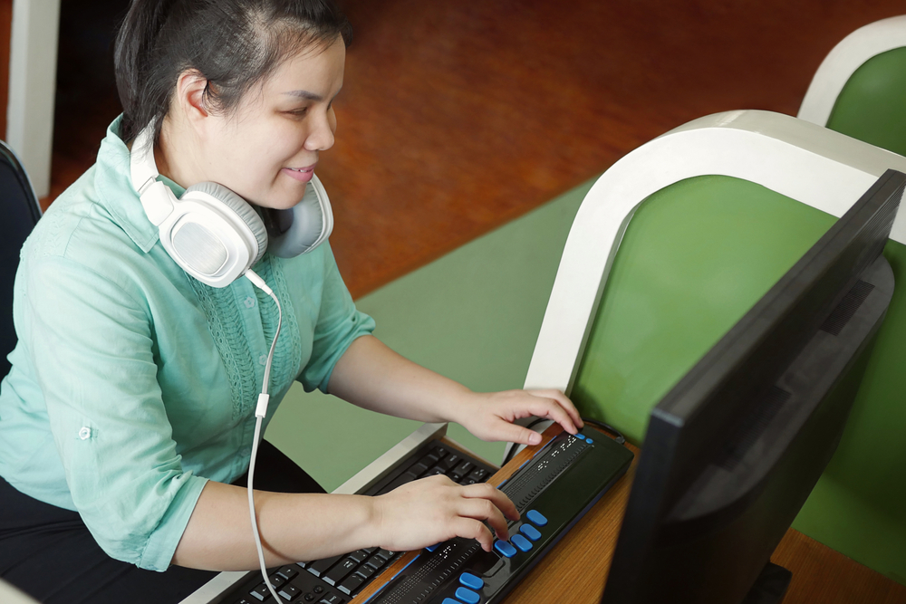 A woman with headphones using computer with a refreshable braille display